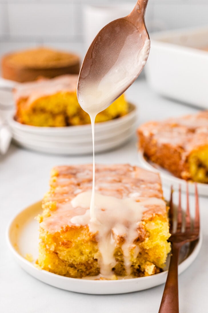 Honey bun cake on a white serving plate with a fork next to it. And a spoon above it drizzling on the topping.