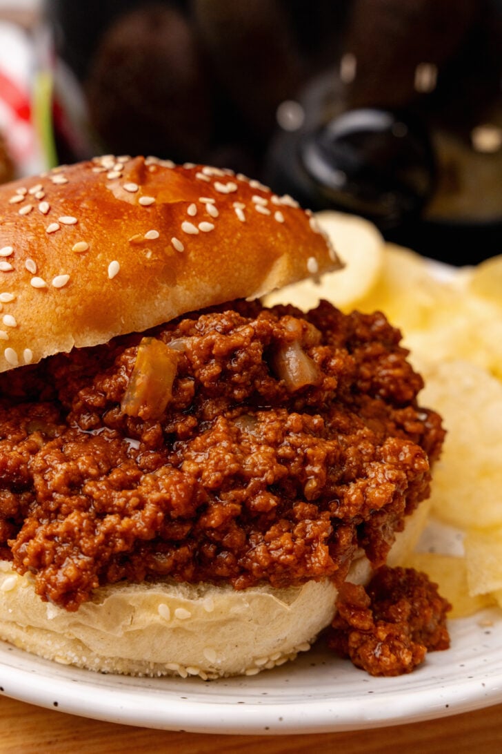 Homemade sloppy joes on a bun next to potato chips sitting on a white plate.