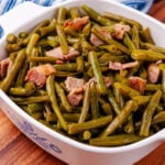 Southern style green beans and perfectly cooked chopped up bacon all sitting inside of a square serving dish sitting on a blue and white kitchen towel.