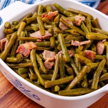 Southern style green beans and perfectly cooked chopped up bacon all sitting inside of a square serving dish sitting on a blue and white kitchen towel.