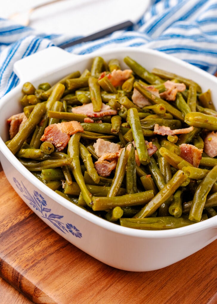 Southern style green beans and perfectly cooked chopped up bacon all sitting inside of a square serving dish sitting on a blue and white kitchen towel.