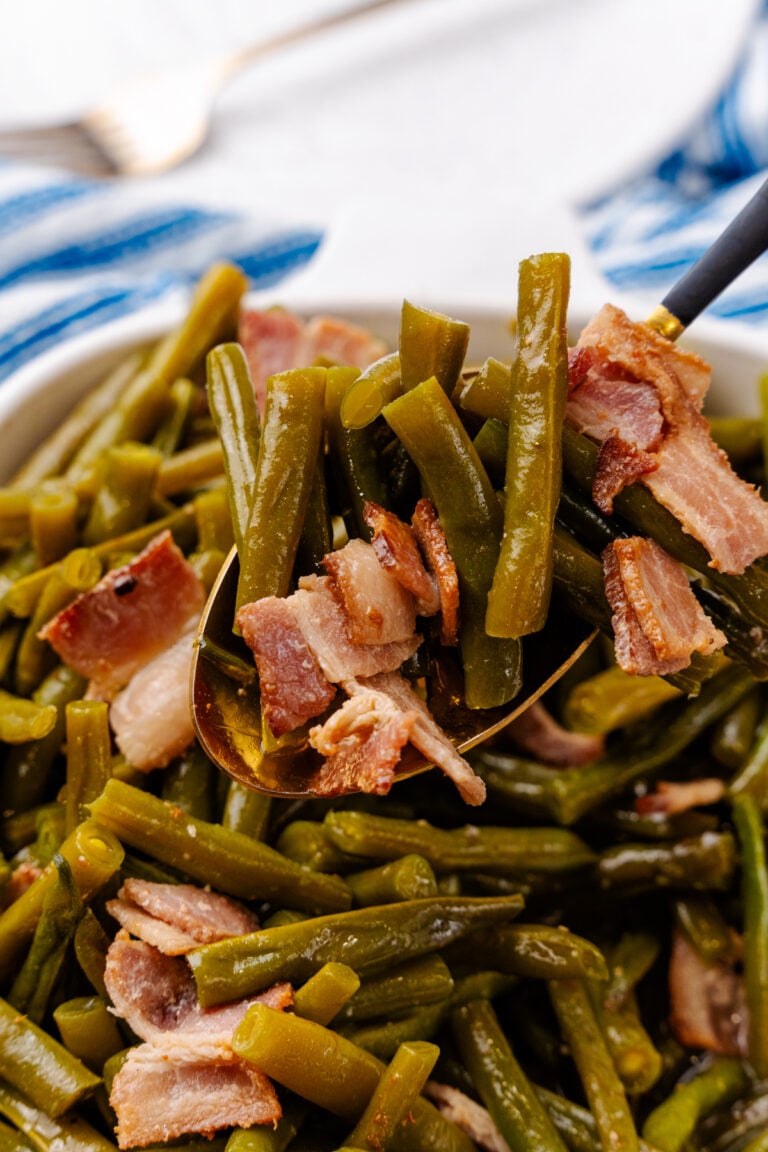 Southern style green beans being scooped out of a white serving dish. You can see the bacon hanging off the sides as-well.