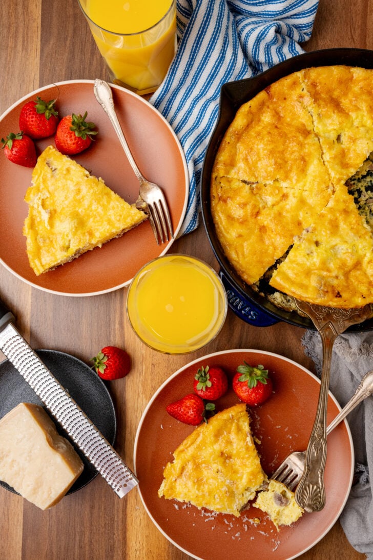 Cast iron pan with frittata being scooped out of it. With two made plates with slices of frittata on it with slices of strawberries.
