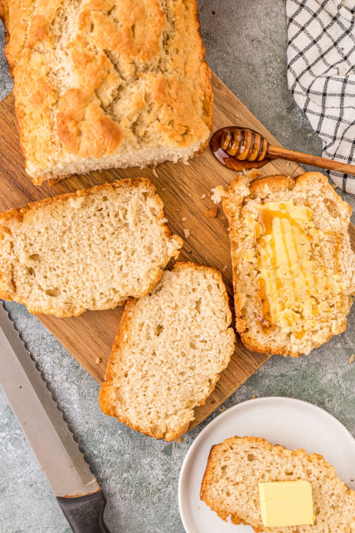 3 slices of beer bread with on having a spread of butter and honey on it. Sitting on a cutting board with a dowel of honey on the side and another slice of beer bread sitting on a white plate.