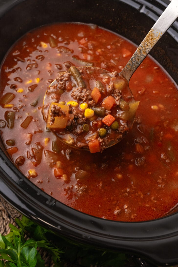 Silver ladle with vegetable beef soup served from a black crock pot.