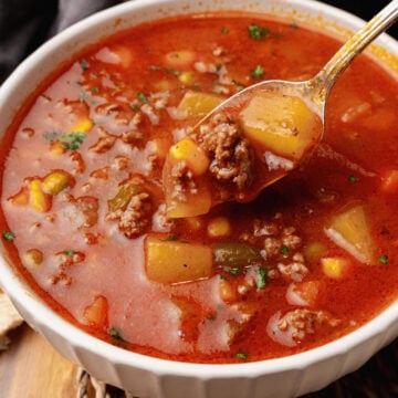 Vegetable beef soup in red tomato broth in a white bowl resting on a woven placemat.