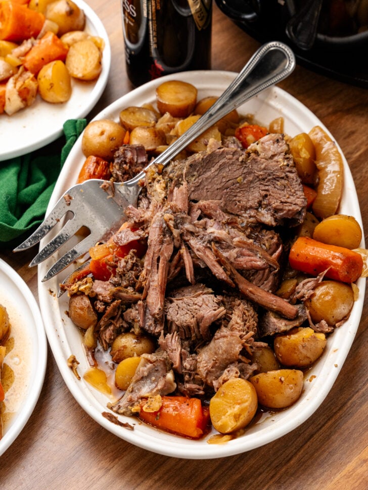 Slicked Irish pot roast with potatoes, carrots, and onions on a white platter served with a metal fork.