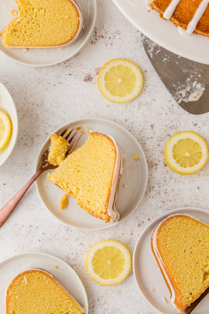 Sliced lemon pound cake on white plate served with a metal fork, on a white counter with lemon slices.