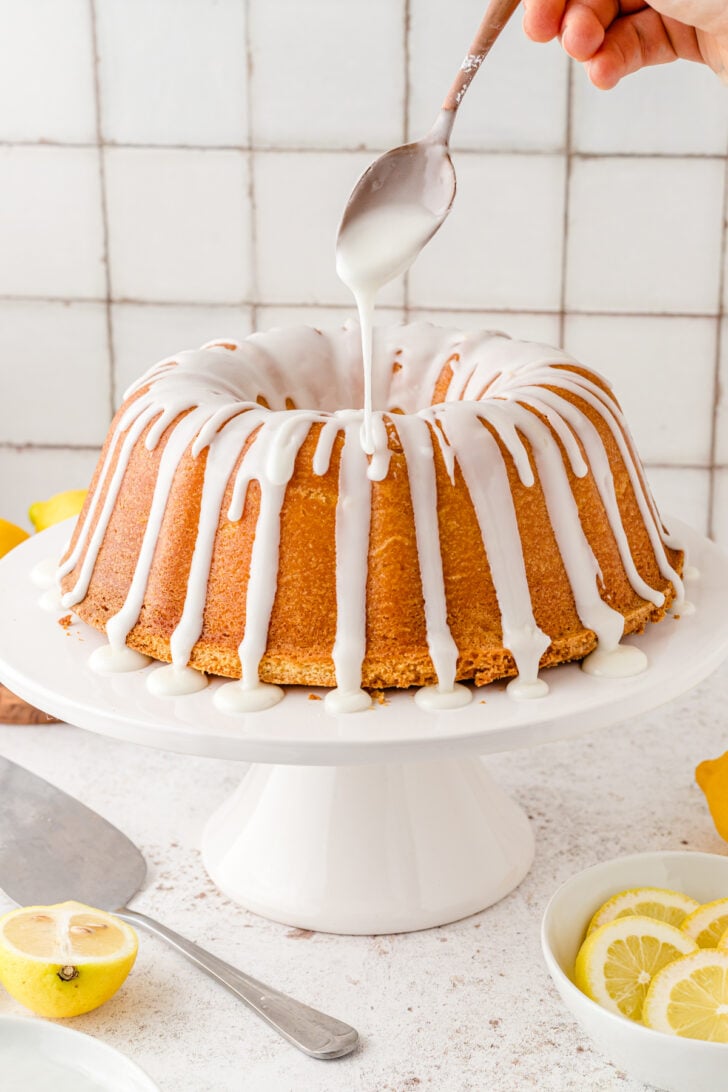 Lemon pound cake on a white cake stand being drizzled with lemon glaze from a metal spoon.