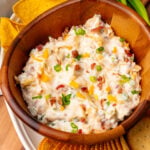 Shredded cheddar cheese green onion and candied bacon all showing on top of a freshly made million dollar dip all put inside a nice wooden serving bowl. with green onion ritz crackers and tortilla chips on the side ready for dipping.