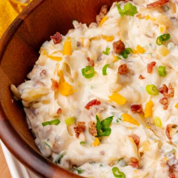 Shredded cheddar cheese green onion and candied bacon all showing on top of a freshly made million dollar dip all put inside a nice wooden serving bowl.