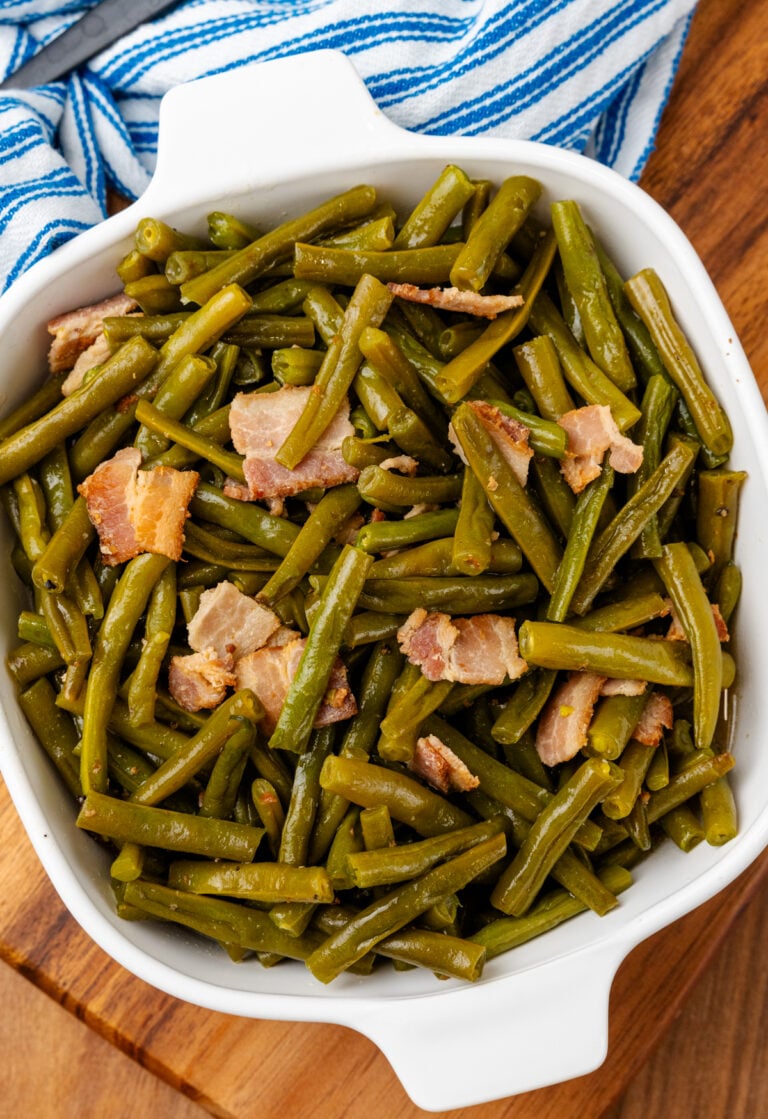 Southern style green beans and perfectly cooked chopped up bacon all sitting inside of a square serving dish sitting on a blue and white kitchen towel.