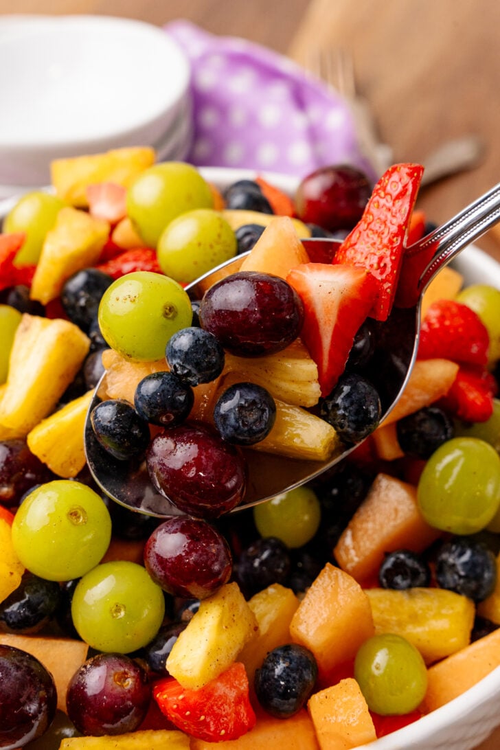 Metal spoonful of fresh fruit salad from a white bowl tossed with orange juice cinnamon dressing.