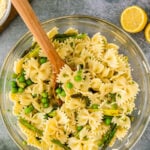 Overhead shot of lemon pasta salad. Green peas bow tie pasta and asparagus in a clear glass bowl with a wooden spoon dipping in.