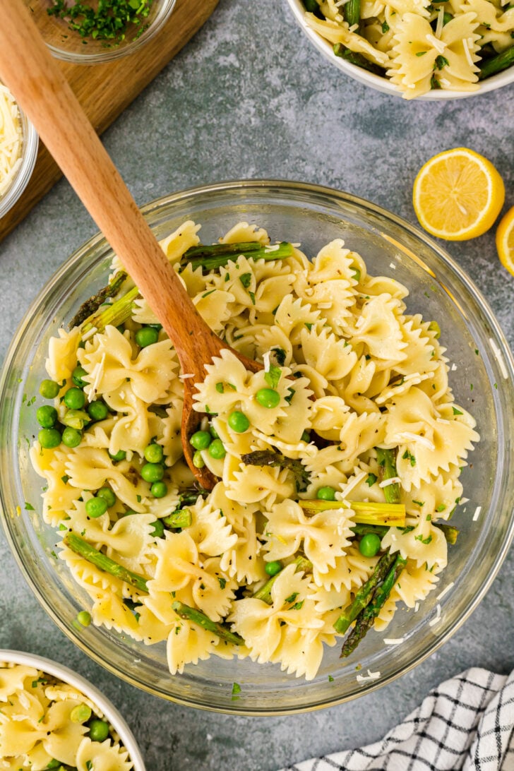 Overhead shot of lemon pasta salad. Green peas bow tie pasta and asparagus in a clear glass bowl with a wooden spoon dipping in.