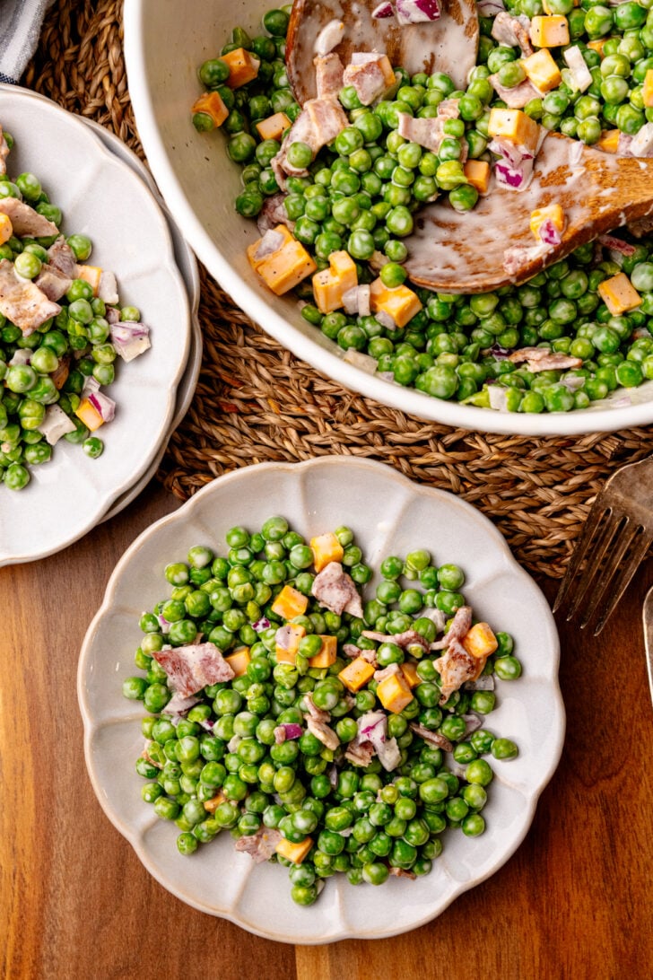 Green peas with cooked bacon, cubed cheddar cheese, and chopped red onion mixed with a creamy dressing served on small white plates.