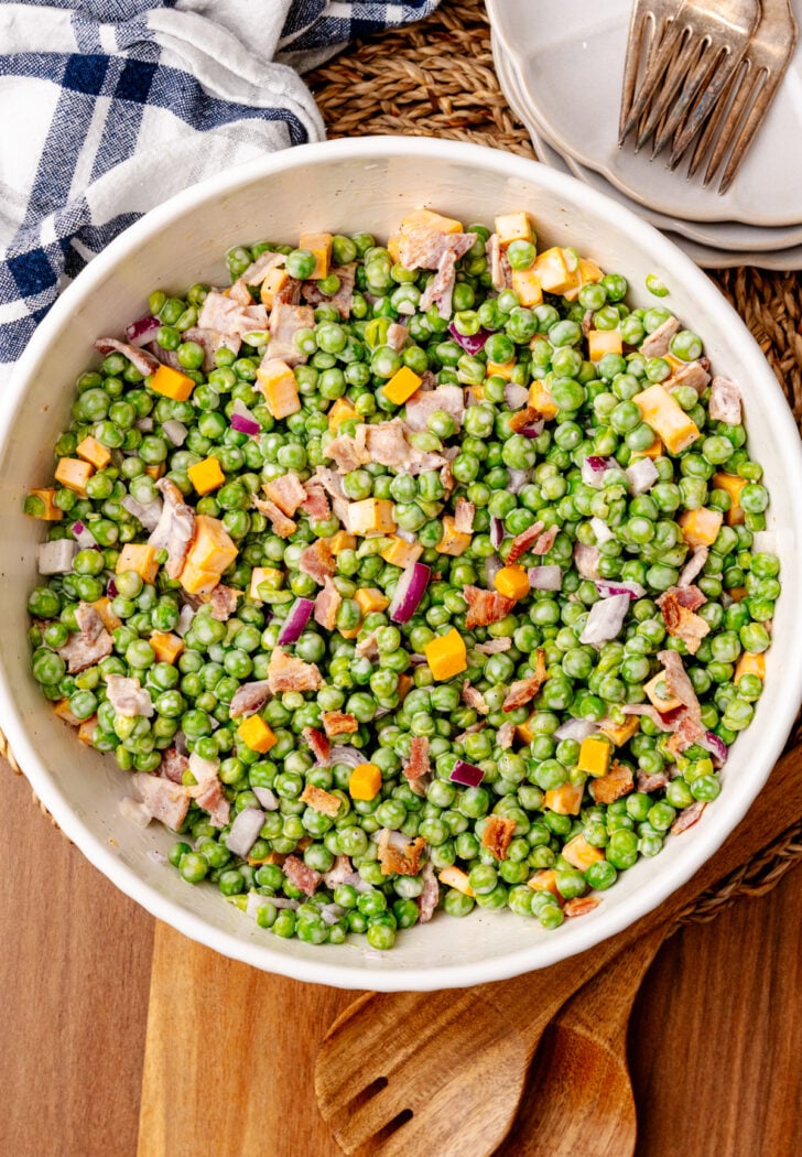 English pea salad 7 in white bowl with bacon, cheese, and peppers on wooden board with plates and napkin