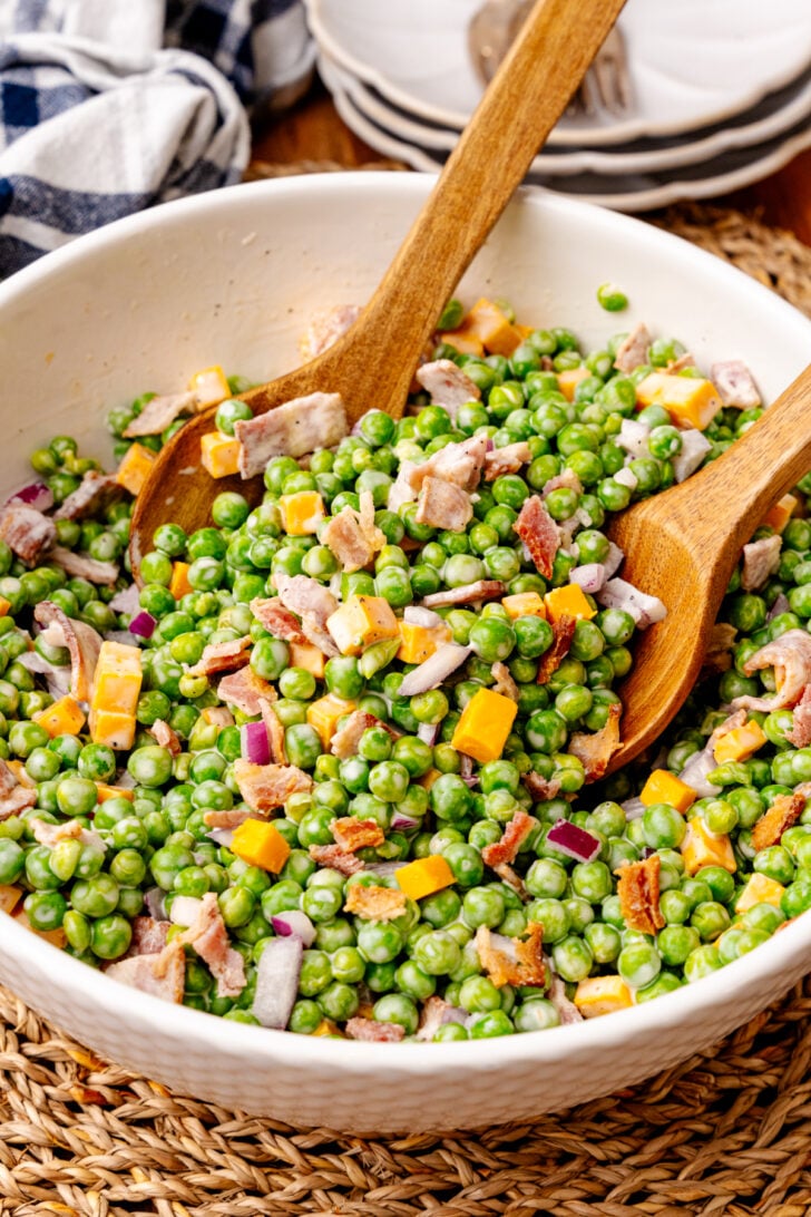 Green peas salad with cheddar cheese, bacon and red onion in a white bowl with wooden serving spoons.