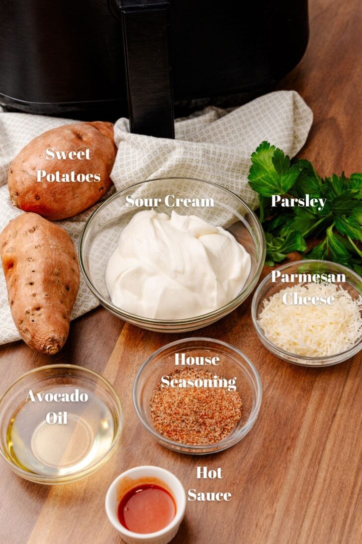 Ingredients on a wooden surface include sweet potatoes, a bowl of sour cream, parsley, grated parmesan cheese, house seasoning, avocado oil, and hot sauce. A kitchen towel and part of an air fryer are visible in the background.