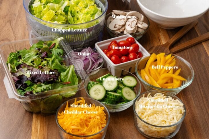 Lettuce, tomatoes, mushrooms, onions, cucumbers, cheese, and bell peppers in containers on a wooden counter.