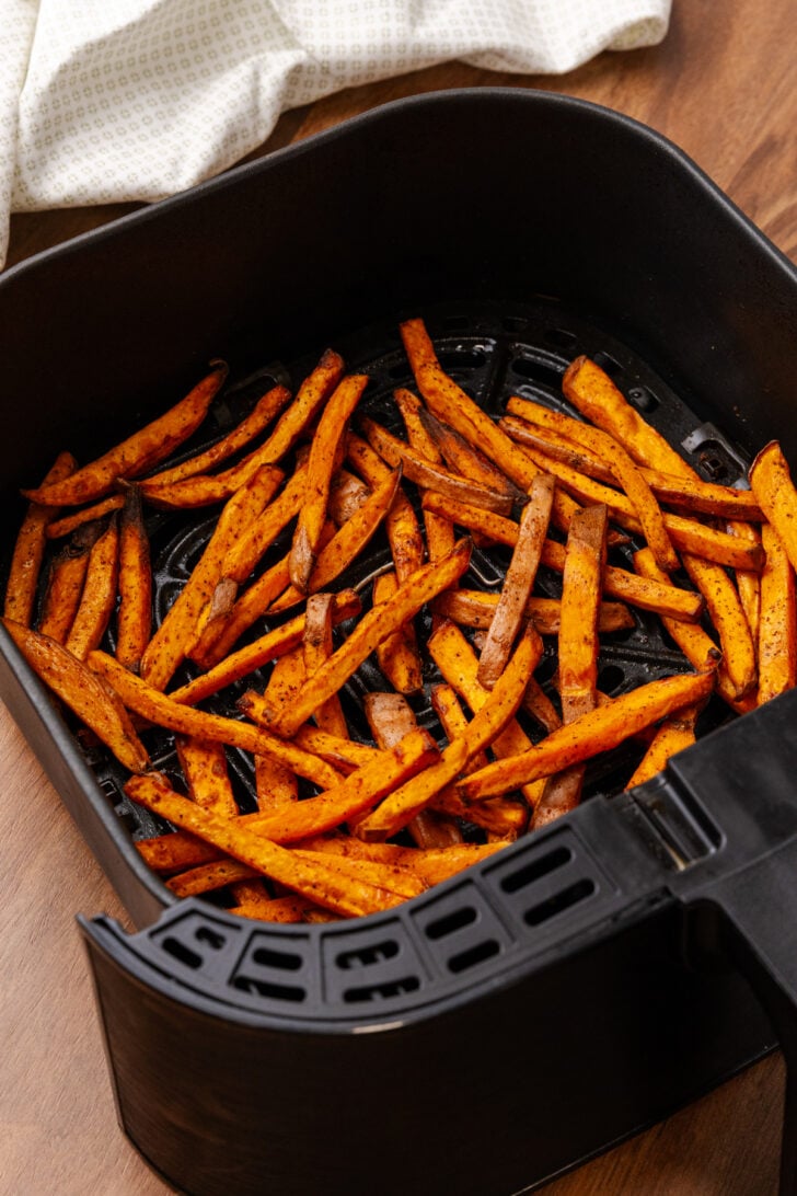 Crispy sweet potato fries are in an air fryer basket, seasoned and evenly spread out. A white cloth is partially visible in the background on a wooden surface.