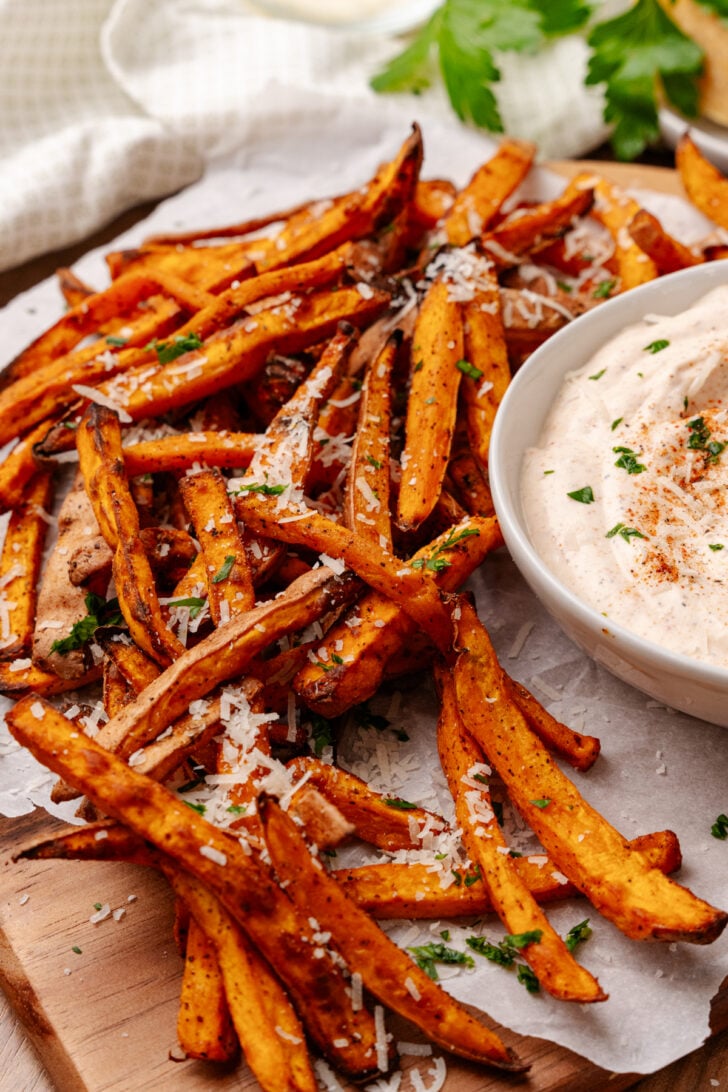 A serving of crispy sweet potato fries garnished with grated cheese and herbs is placed on a wooden platter alongside a bowl of creamy dip, sprinkled with spices and garnished with herbs. Fresh green parsley is visible in the background.