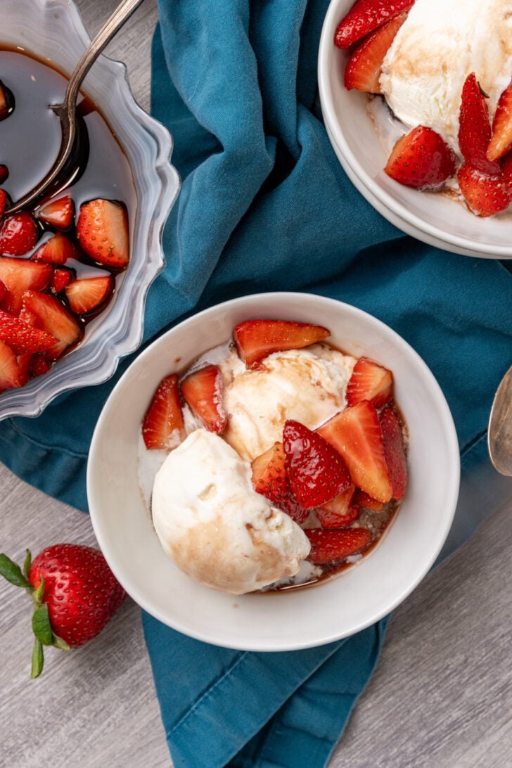 Overhead view of balsamic strawberries with vanilla ice cream in white bowls on teal linen, gray wood surface
