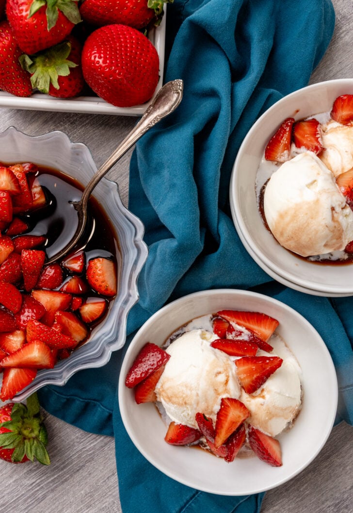 Overhead view of two white bowls with vanilla ice cream topped with balsamic strawberries, alongside a gray dish of macerated berries and fr
