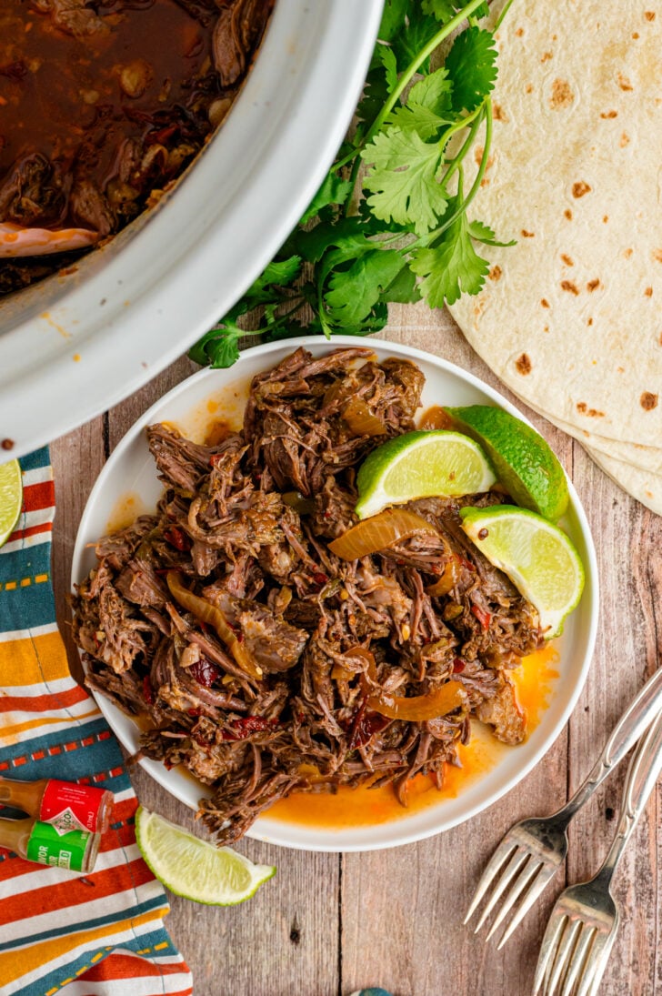 White plate with shredded Mexican beef served with lime wedges and a crock pot with additional beef for serving.