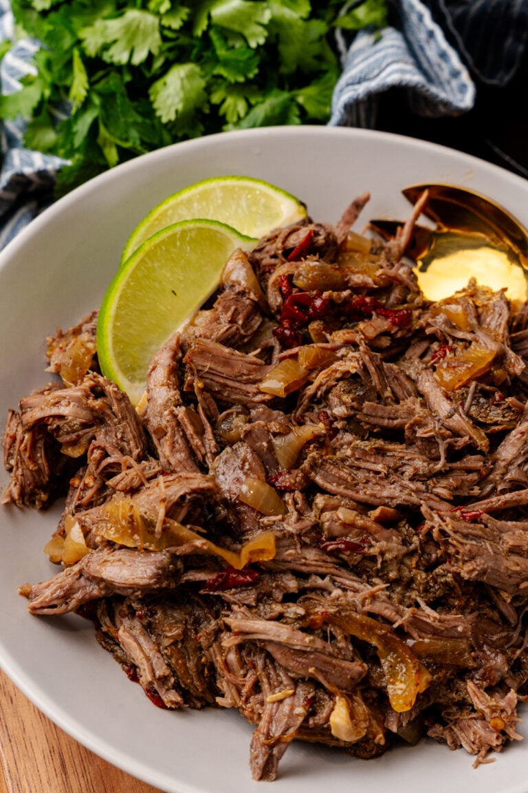 Shredded cp mexican beef 7 on white plate with lime wedges, onions, cilantro garnish, and blue striped cloth