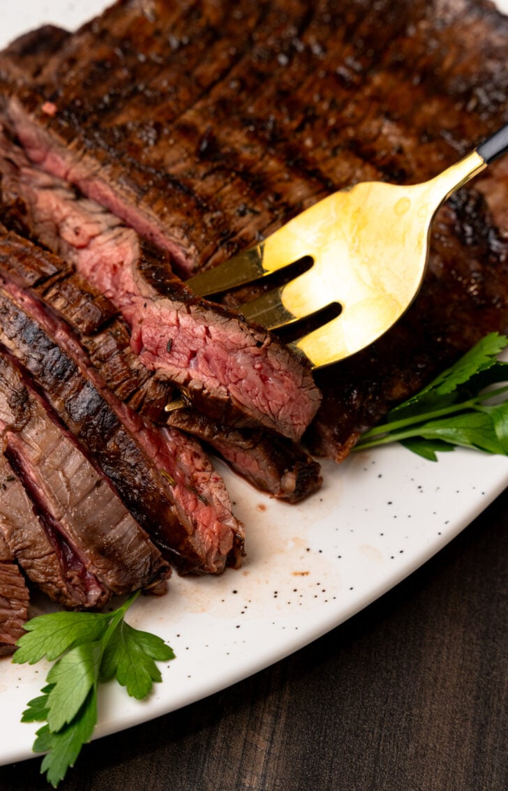Grilled flank steak, sliced into strips and served on a white plate with a gold fork.