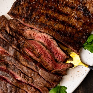 Sliced medium-rare flank steak on a white speckled plate, garnished with fresh parsley and served with mustard