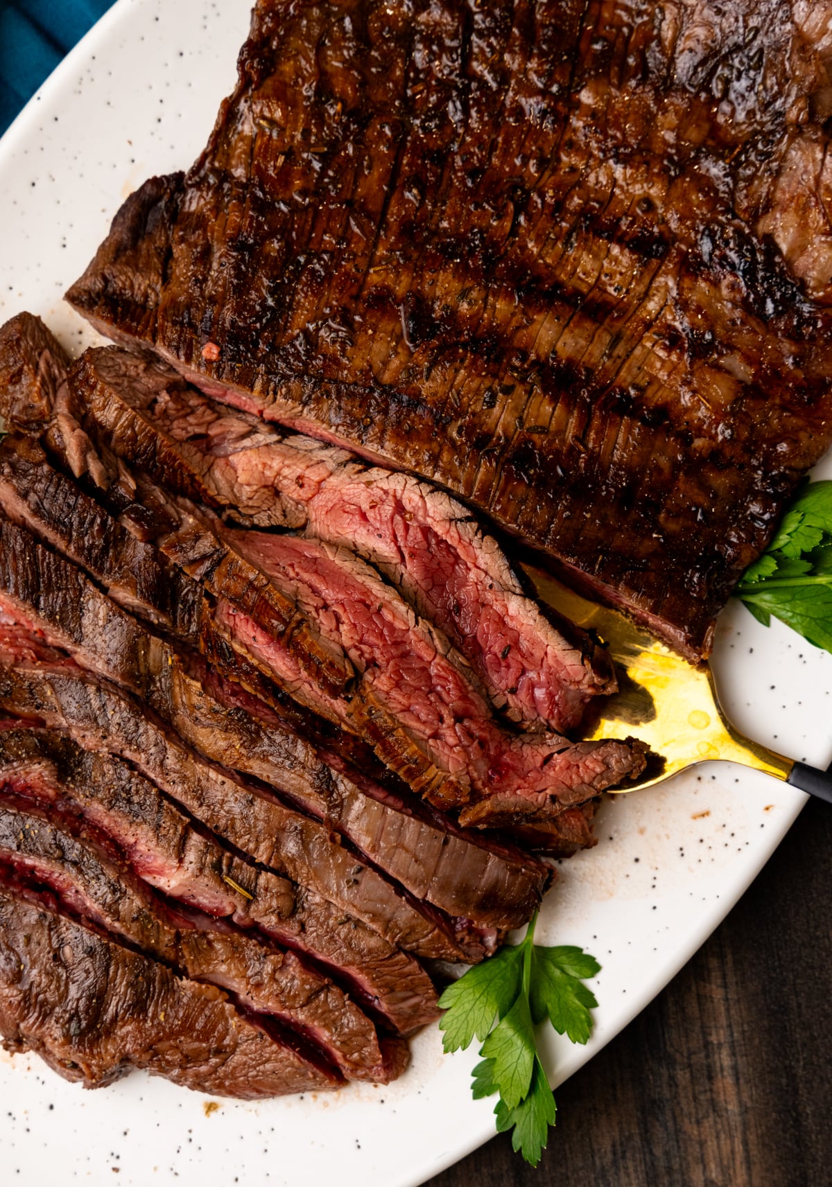 Sliced medium-rare flank steak on a white speckled plate, garnished with fresh parsley and served with mustard