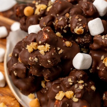 Homemade rocky road clusters topped with mini marshmallows and crushed graham crackers on a white plate with wooden surface