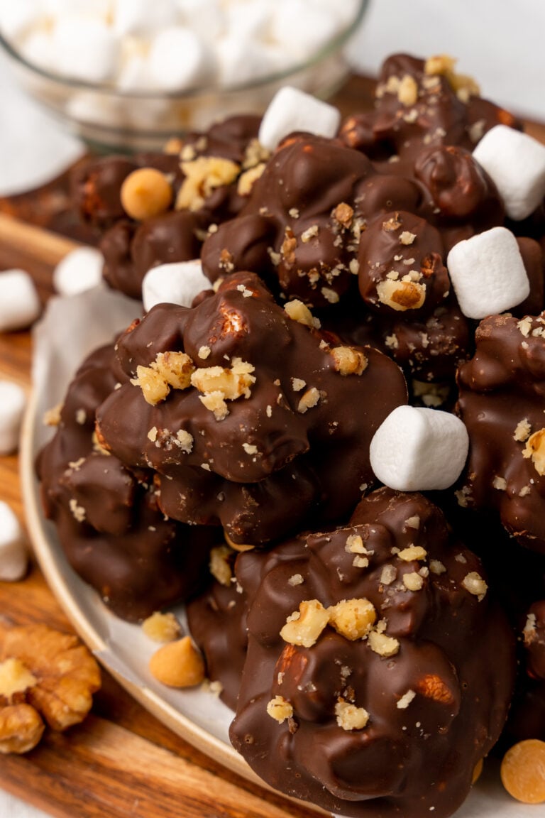 Homemade rocky road clusters topped with mini marshmallows and crushed graham crackers on a white plate with wooden surface