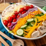 Colorful salad for a crowd 3 1 arranged on white plate with shredded cheese, cherry tomatoes, mushrooms, cucumbers, yellow peppers, and purp