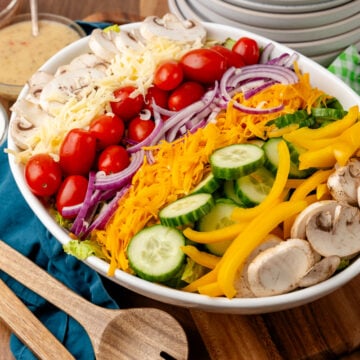 Colorful salad for a crowd 3 1 arranged on white plate with shredded cheese, cherry tomatoes, mushrooms, cucumbers, yellow peppers, and purp