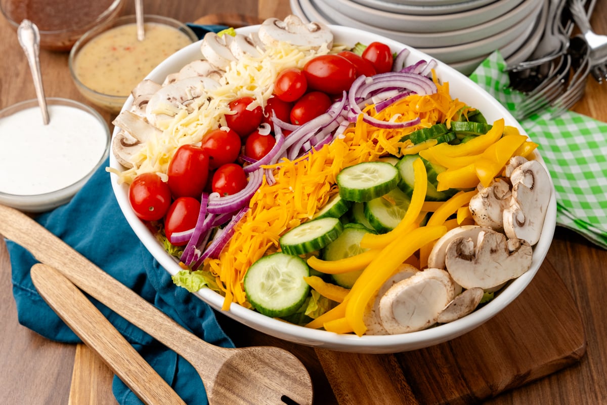 Colorful salad for a crowd 3 1 arranged on white plate with shredded cheese, cherry tomatoes, mushrooms, cucumbers, yellow peppers, and purp