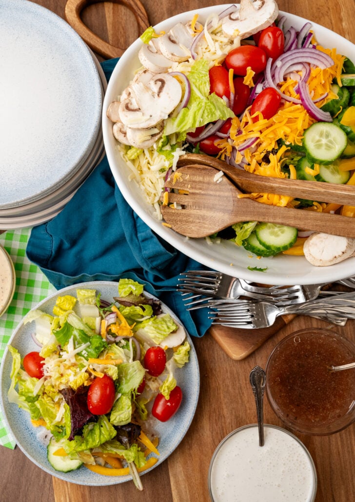 Large white salad bowl with wooden serving tools and a small plate with green salad with tomatoes and cheese.