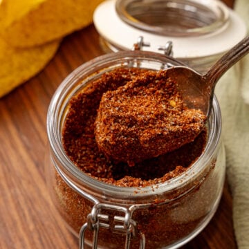 Homemade taco seasoning 4 in glass jar with spoon, yellow peppers and empty jar in background on wooden surface