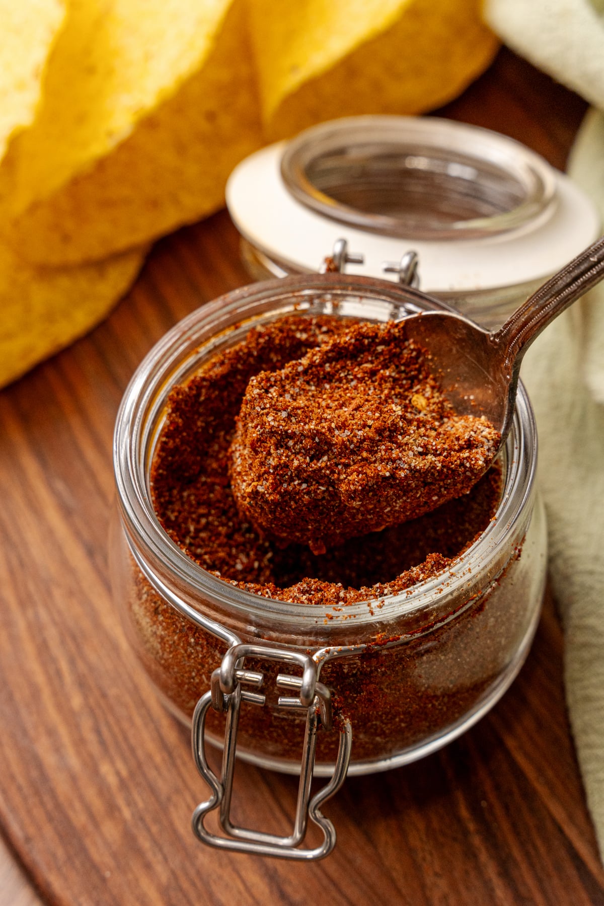 Homemade taco seasoning 4 in glass jar with spoon, yellow peppers and empty jar in background on wooden surface