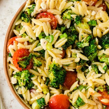 Broccoli orzo salad with cherry tomatoes and green onions in a speckled brown bowl on a light marble surface