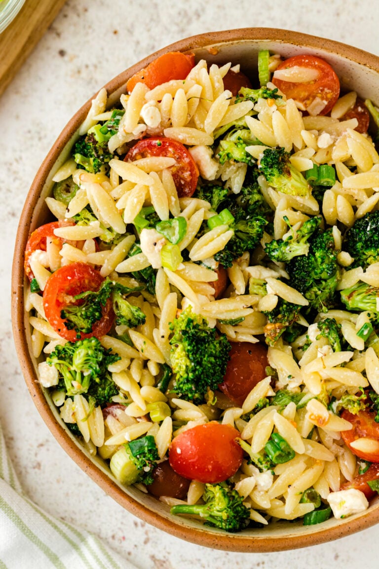 Broccoli orzo salad with cherry tomatoes and green onions in a speckled brown bowl on a light marble surface