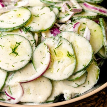 Creamy cucumber salad with sliced cucumbers, red onion, fresh dill, and black pepper in a ceramic bowl on a woven placemat
