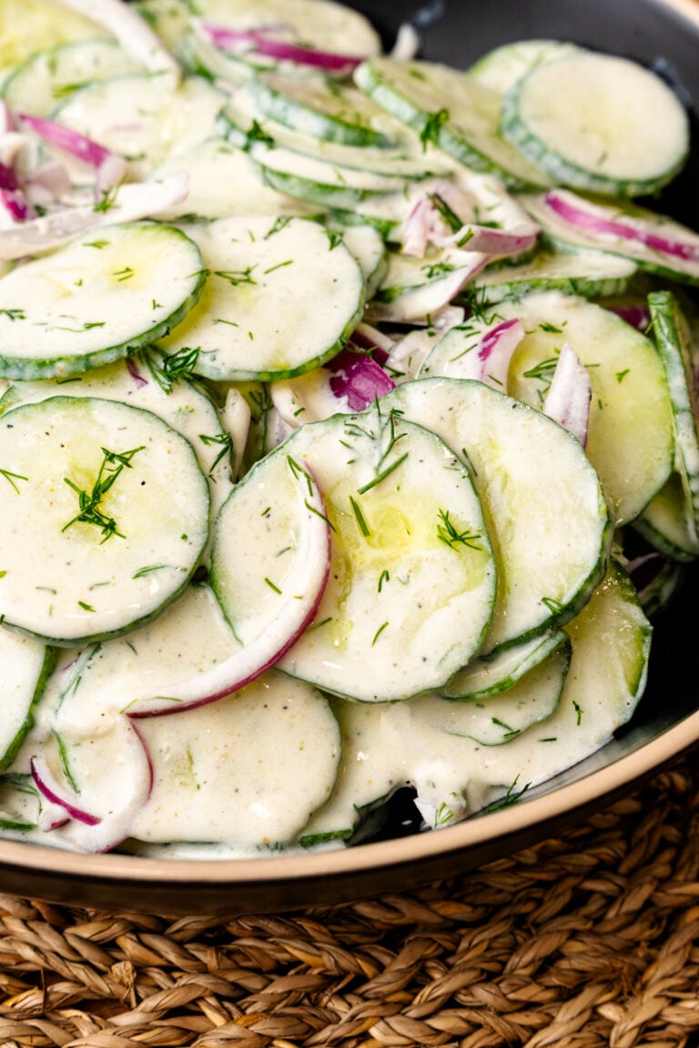 Creamy cucumber salad with sliced cucumbers, red onion, fresh dill, and black pepper in a ceramic bowl on a woven placemat