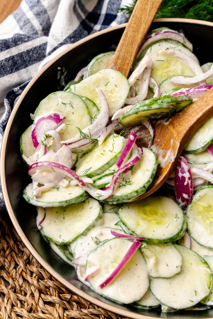 Mixing bowl with sliced green cucumbers, red onions in a creamy dressing mixed with wooden utensils.