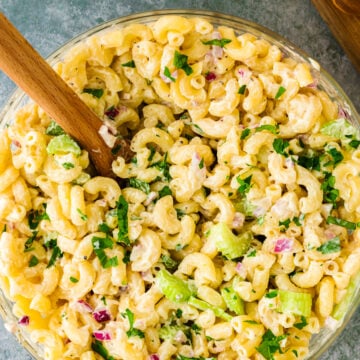 Overhead view of creamy macaroni salad 9 in a glass bowl with wooden spoon, topped with fresh parsley, celery, and diced ham
