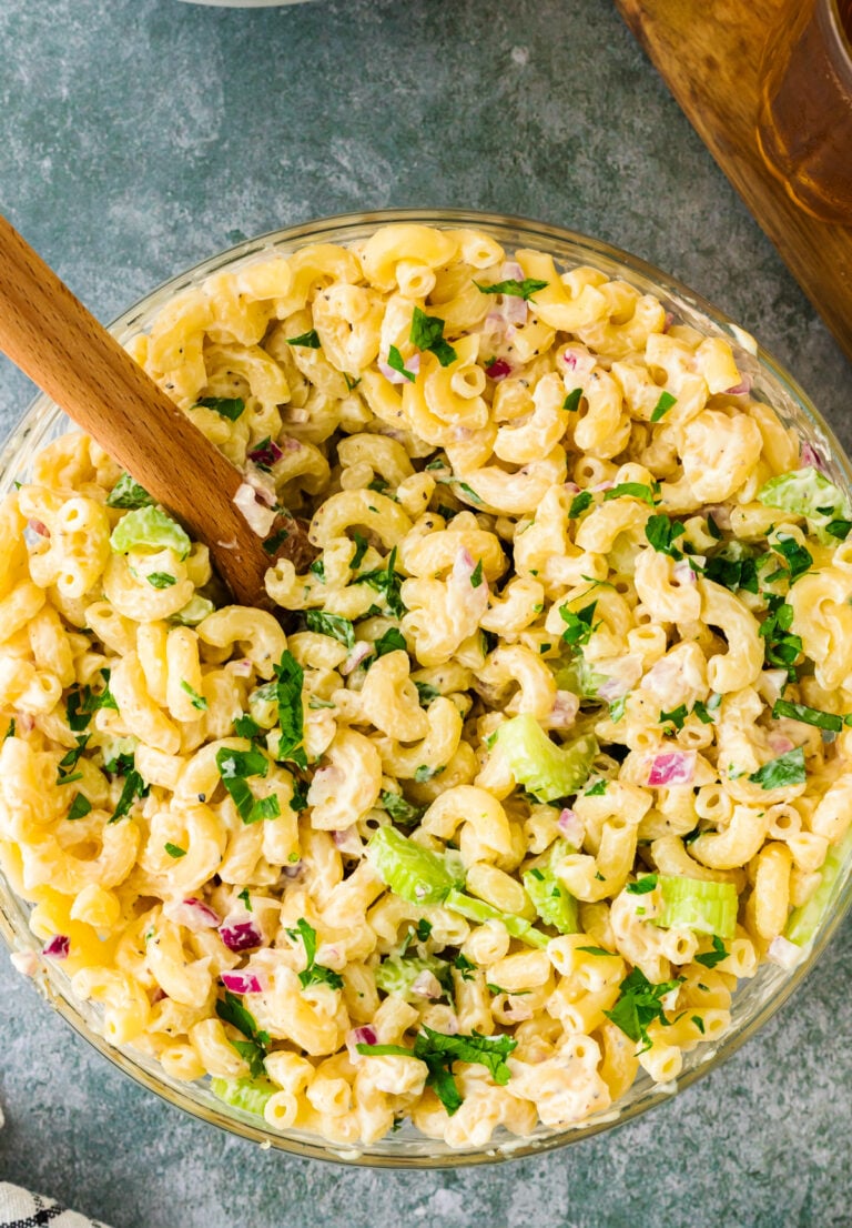 Overhead view of creamy macaroni salad 9 in a glass bowl with wooden spoon, topped with fresh parsley, celery, and diced ham