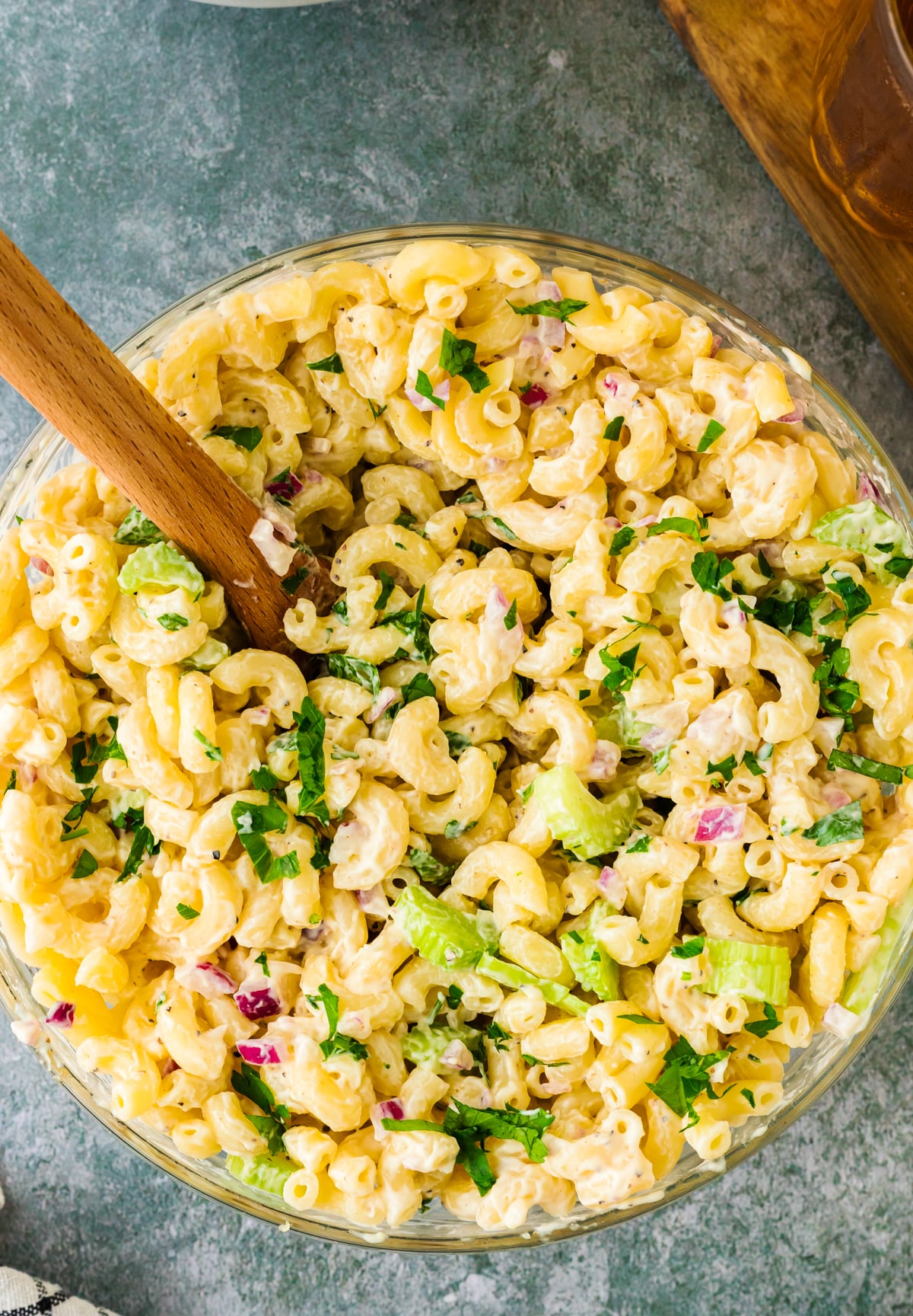 Overhead view of creamy macaroni salad 9 in a glass bowl with wooden spoon, topped with fresh parsley, celery, and diced ham