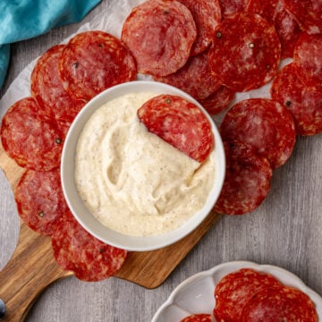 Crispy salami chips arranged around a white bowl of creamy dip on a wooden board with parchment paper and a blue napkin